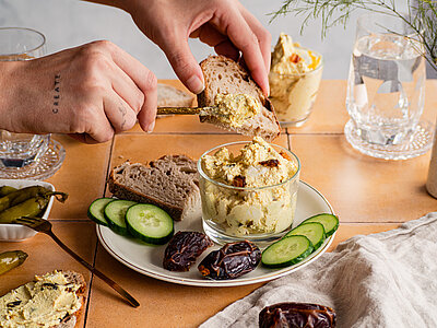 Tofu-Curry-Dattel-Dip Hellbraune Creme mit Datteln, Gurkenscheiben und Brotscheiben auf einem Teller mit Tischdekoration.