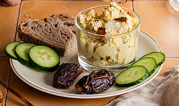 Tofu-Curry-Dattel-Dip Hellbraune Creme mit Datteln, Gurkenscheiben und Brotscheiben auf einem Teller mit Tischdekoration.