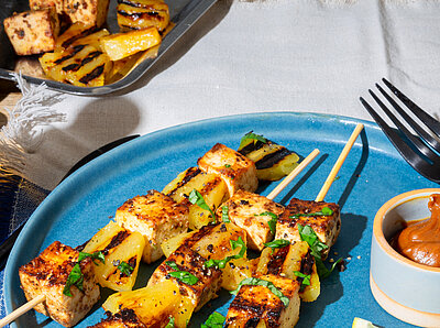 Ananas-Spiesse Zitronen-Knoblauch-Marinade Drei Tofu-Spiesse mit Dip und Früchtedekoration auf blau-weißem Tischtuch.