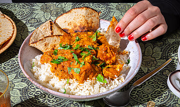 Tikka Masala Tofuwürfel mit roter Soße auf Reis mit dünnem Fladenbrot auf einem Teller mit Tischdekoration. Eine Hand am Teller.