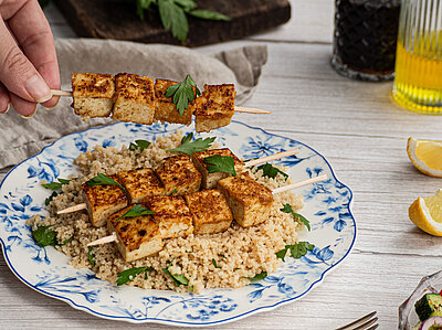 Harissa Tofu Spiesse mit Couscous Tofu-Spiesse auf Couscous auf einem Teller mit buntem Gemüse und Tischdekoration.