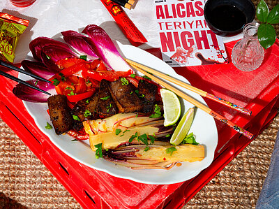 Teriyaki Tofu mit Gemüse Dunkle Tofuscheiben an Gemüse auf einem Teller mit Tischdekoration. Jemand hält Essstäbchen.