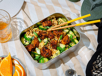 Asia-Nudelsalat mit Orangen-Gojuchang-Tofu Nudelsalat mit Gurken und gewürztem Tofu in einer Metallschüssel auf einem Tischtuch mit Deko.