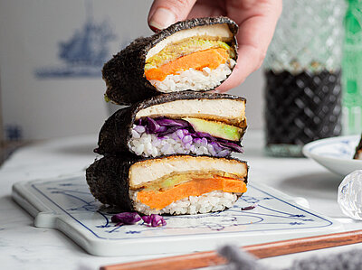 Sushi-Sandvich Eine Hand hält Sushi in 3 Algenblättern mit Tofu, Reis, Rotkohl und Süßkartoffel auf einem Brett mit Tischdekoration.