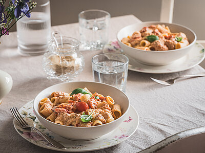 Cremige Veta-Pasta Pasta mit Soße, Tomaten und Basilikum auf Tellern und Wassergläser auf einem Tischtuch mit Dekoration.