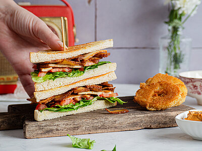 Bacon-Sandvich Zwei Toastscheiben belegt mit Bacon auf einem Holzbrett und Dekoration wird von einer Hand gehalten.