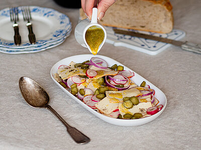 Räucher-Tofu-Salat Dünne Scheiben Tofu mit Zwiebeln, Gurken und Radieschen in einer Schale, jemand gießt gelben Dressing darauf, mit Tischdekoration.