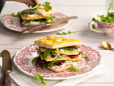 Tofu-Waffle Vier übereinanderliegende Waffeln, dazwischen Tofu, getrocktete Tomaten und Salat aufeinem Teller mit Tischdekoration.