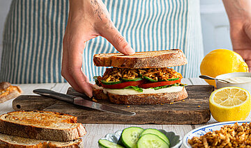 Räuchertofu Protein Sandwich Zwischen 2 Graubrotscheiben Tofu-Stückchen und Salat auf einem Holzbrett und Dekoration.
