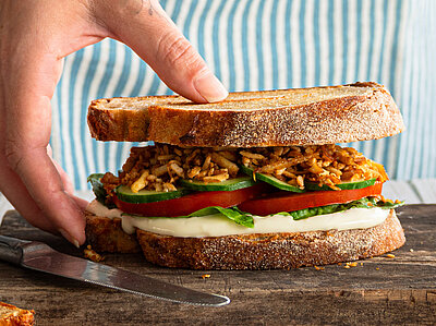 Räuchertofu Protein Sandwich Zwischen 2 Graubrotscheiben Tofu-Stückchen und Salat auf einem Holzbrett und Dekoration.