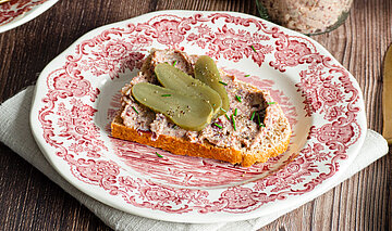 Lebervurst Ein Glas mit einer Tofupaste, daneben ein Brot auf einem Teller mit der Tofupaste und Gurkenscheiben mit Dekoration.